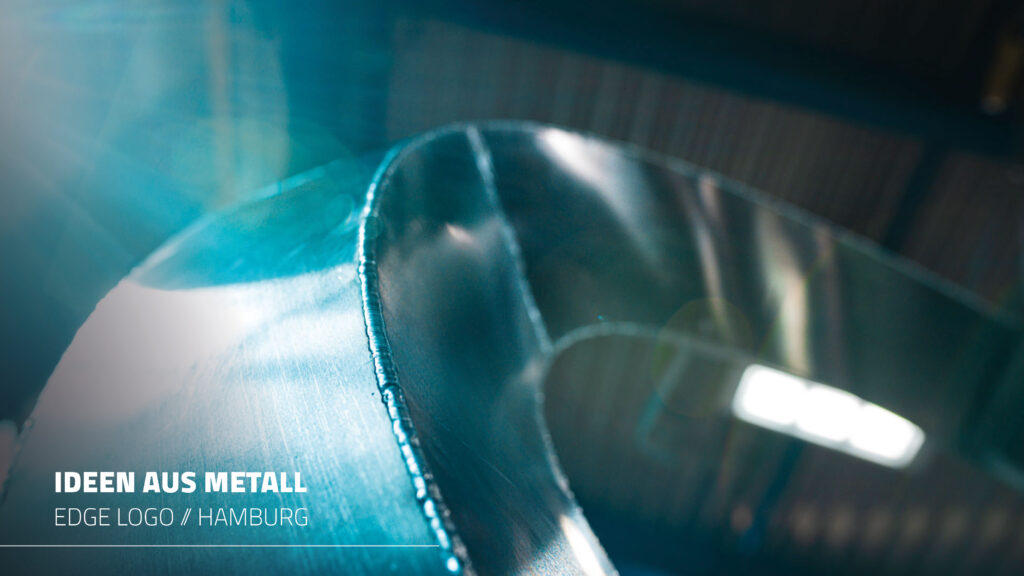 Eine abstrakte Makroaufnahme eines glänzenden Metallobjekts in blauem Farbton und Lichtreflexionen. Die Worte „IDEEN AUS METALL“ und „EDGE LOGO // HAMBURG“ sind im Bild eingeblendet.