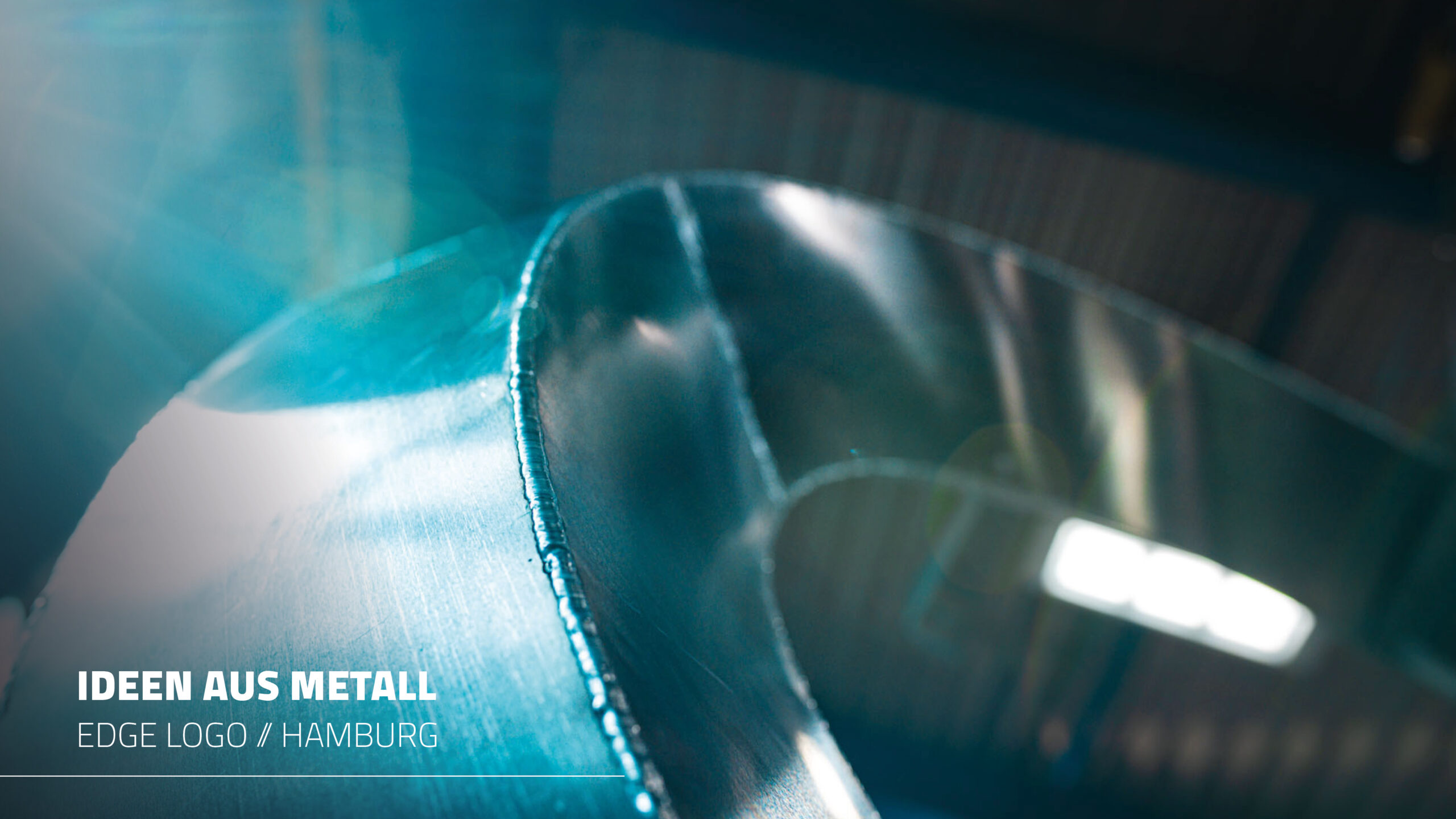 Eine abstrakte Makroaufnahme eines glänzenden Metallobjekts in blauem Farbton und Lichtreflexionen. Die Worte „IDEEN AUS METALL“ und „EDGE LOGO // HAMBURG“ sind im Bild eingeblendet.