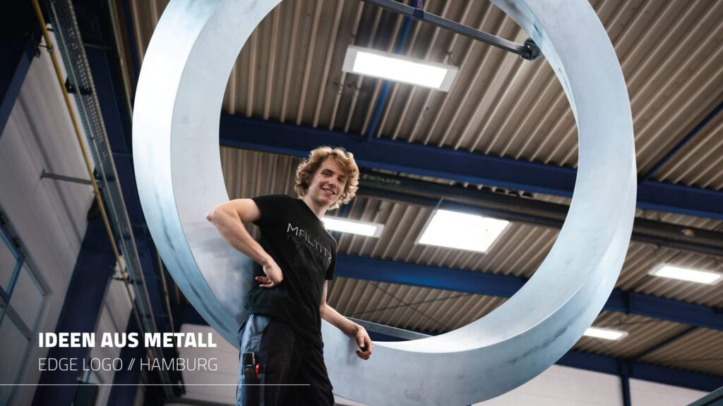 Mitarbeiter Merlin steht lässig angelehnt an das riesige, geschwungene runde Metalllogo, und schaut nach unten in die Kamera