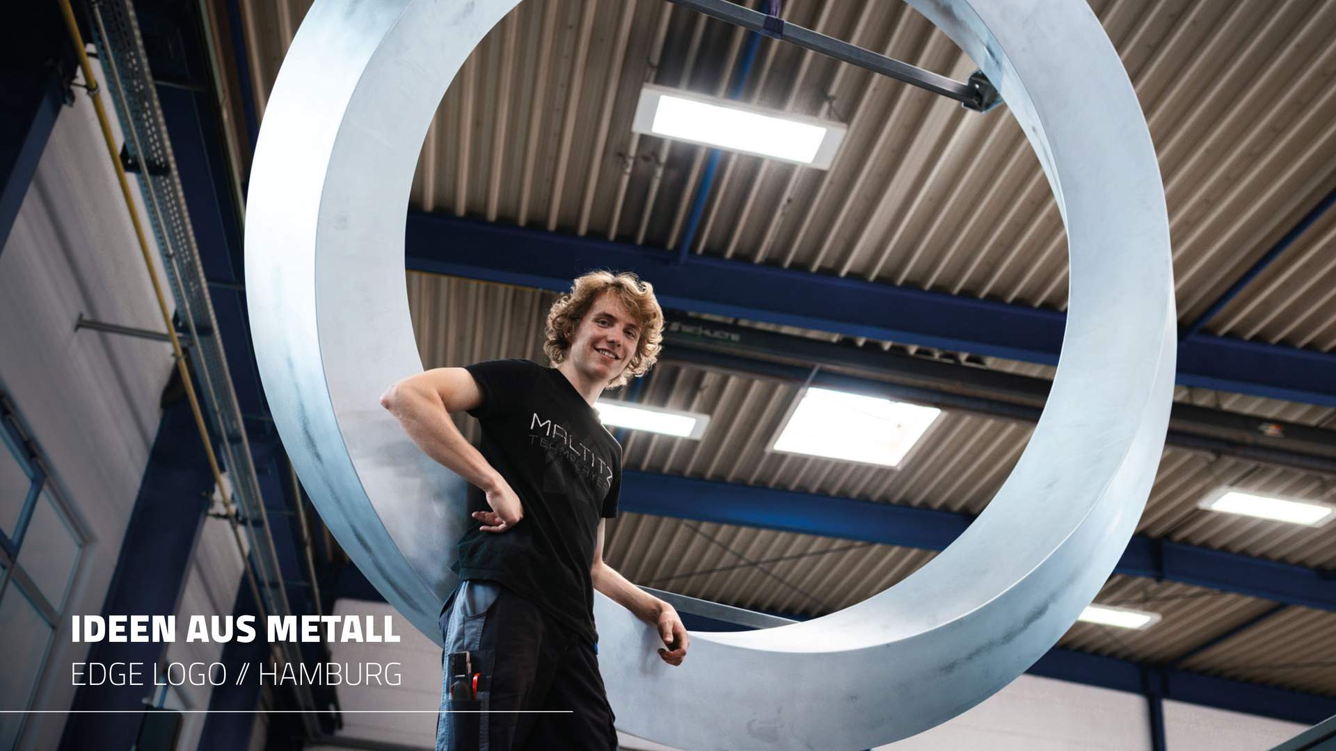 Mitarbeiter Merlin steht lässig angelehnt an das riesige, geschwungene runde Metalllogo, und schaut nach unten in die Kamera