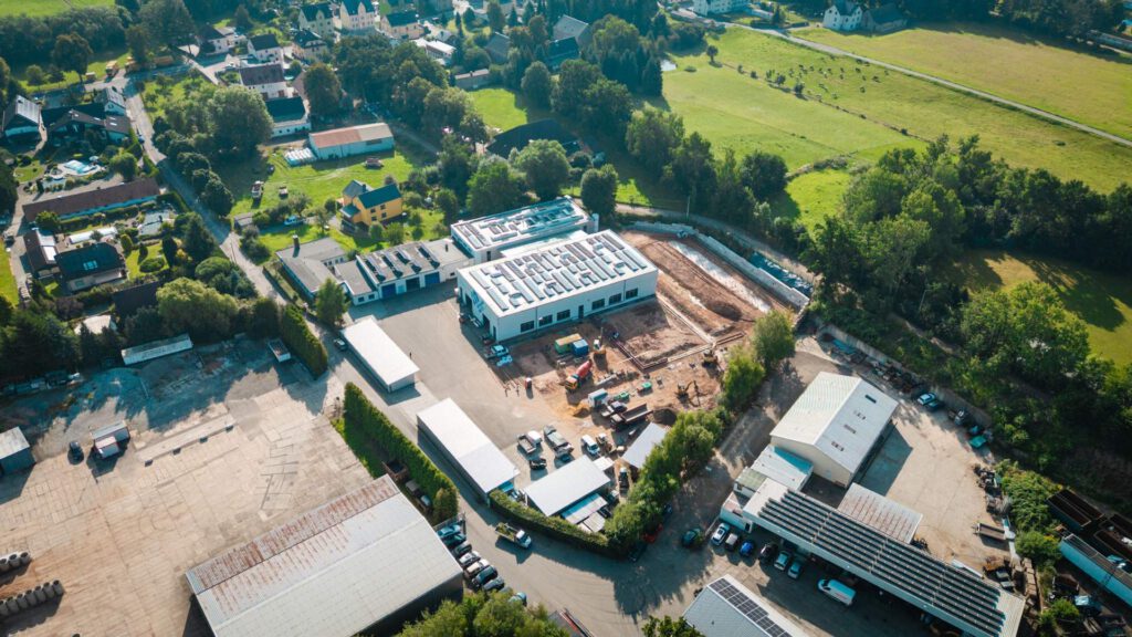 Eine hochwinkelige Luftaufnahme einer großen Baustelle in ländlicher Umgebung, die das MALTITZ-Gebäude sowie die aktuelle Baustelle, Baufahrzeuge und umliegende grüne Felder zeigt.