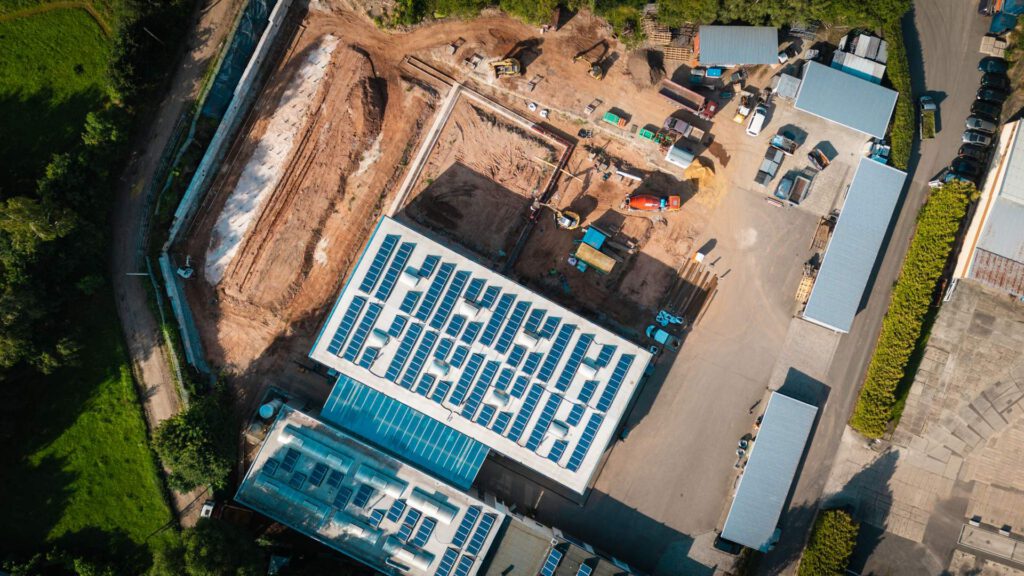 Eine Luftaufnahme von oben auf eine Baustelle, die ein großes Gebäude mit einer umfangreichen Solaranlage auf dem Dach sowie verschiedene Baumaschinen und Materialien auf dem Boden hervorhebt.