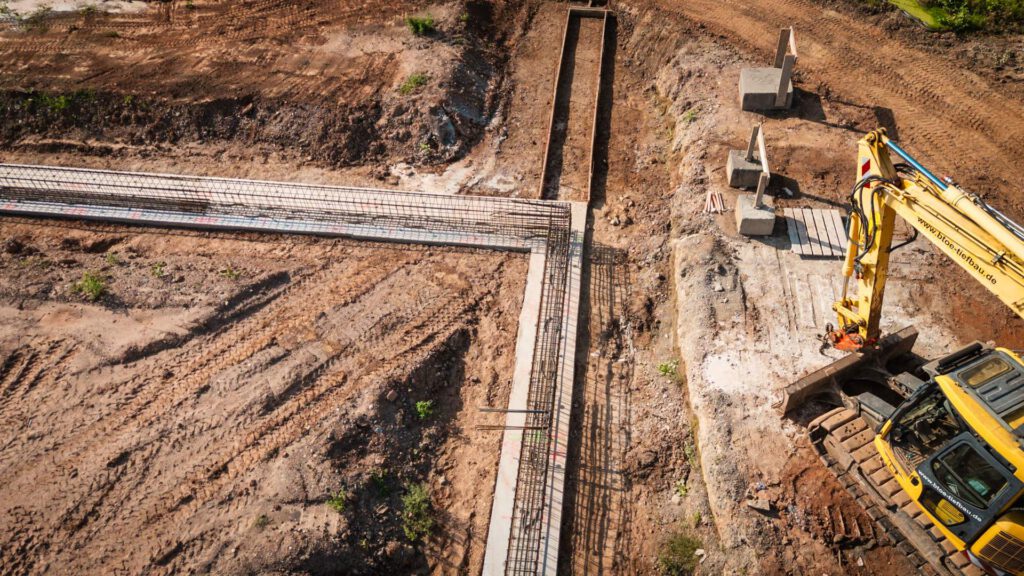 Luftaufnahme einer Baustelle mit einem neu gegossenen Fundament inklusive Stahlbewehrung und einem gelben Bagger.
