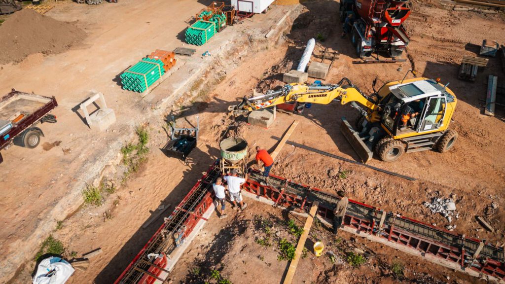 Eine Luftaufnahme von Bauarbeitern, die Beton in eine rote Schalung gießen, um ein Fundament zu erstellen. Ein Liebherr-Bagger und andere Maschinen sind im Hintergrund zu sehen.