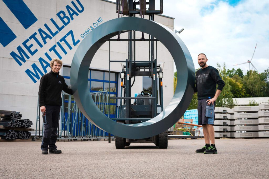 Mitarbeiter Merlin (links) und Daiel (rechts) stehen vor der MALTITZ Werkhalle, das fertige Logo hängt an eine Gabelstapler in der Mitte von ihnen
