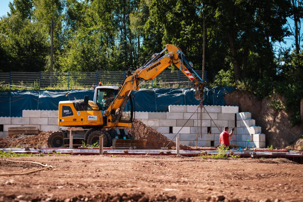 Eine Nahaufnahme auf Bodenebene einer Baustelle, die einen gelben Liebherr-Bagger zeigt, der einen Betonblock anhebt, während ein Bauarbeiter in einem roten Hemd den Block neben einer neu gebauten Stützmauer in Position bringt.