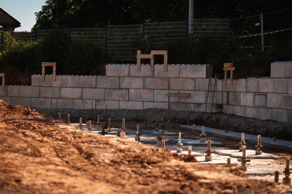 Eine Ansicht auf Bodenebene einer Baustelle, die eine neu gebaute Stützmauer aus Betonblöcken und die Metallbewehrung sowie Ankerbolzen des zukünftigen Fundaments der neuen Werkhalle zeigt.
