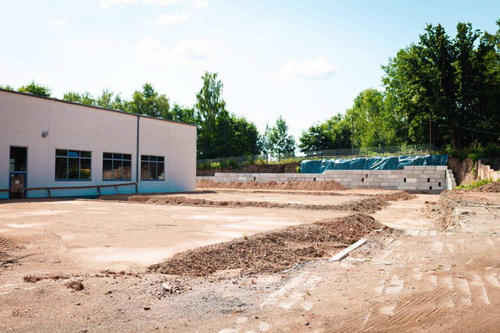 Aufnahme einer Baustelle mit der weißen Werkhalle von Maltitz, freiem Erdbereich und einer Stützmauer aus Betonblöcken im Hintergrund.