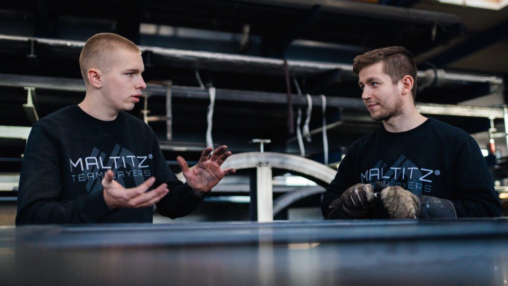 Zwei junge Männer in schwarzen MALTITZ®-Teamplayers-Pullover besprechen ihre Arbeit in der Produktionshalle.