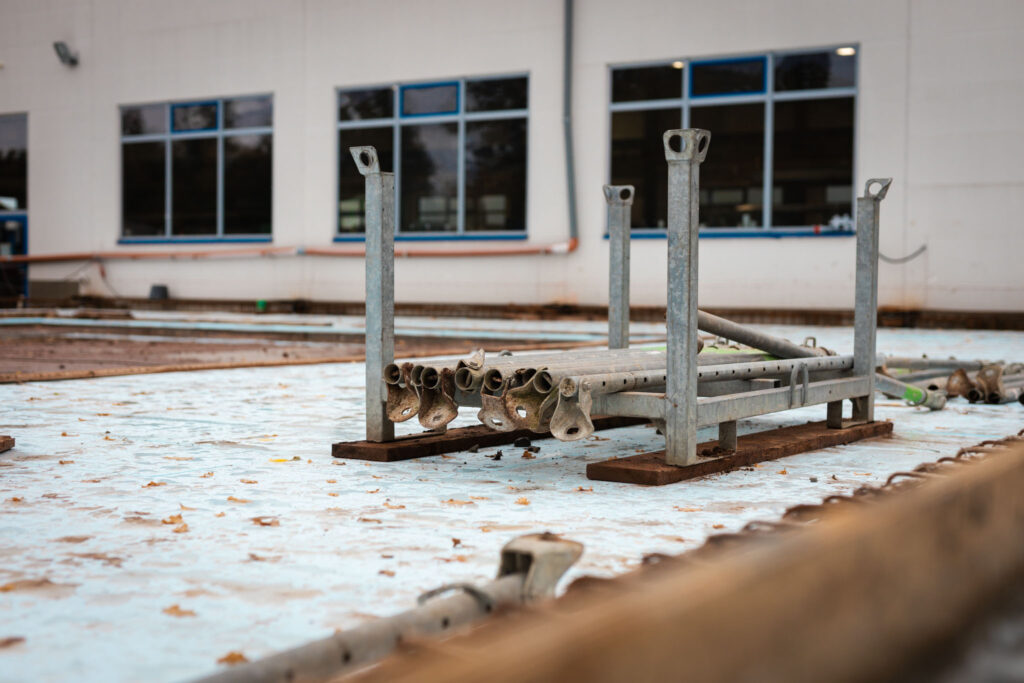 Nahaufnahme von gestapelten Gerüstrohren und Metallständern auf einer mit Geröll bedeckten Baufläche vor einem Gebäude.