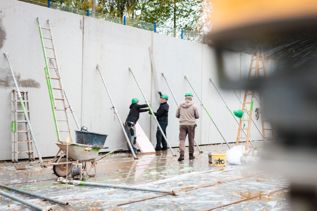 Drei Bauarbeiter montieren Stützstreben an einer langen Betonwand auf einer nassen Baustellenfläche, mit Leitern und Baugeräten.