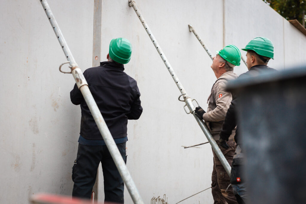 Drei Bauarbeiter in Schutzhelmen stützen eine hohe Betonwand mit Stützstreben auf einer Baustelle ab.