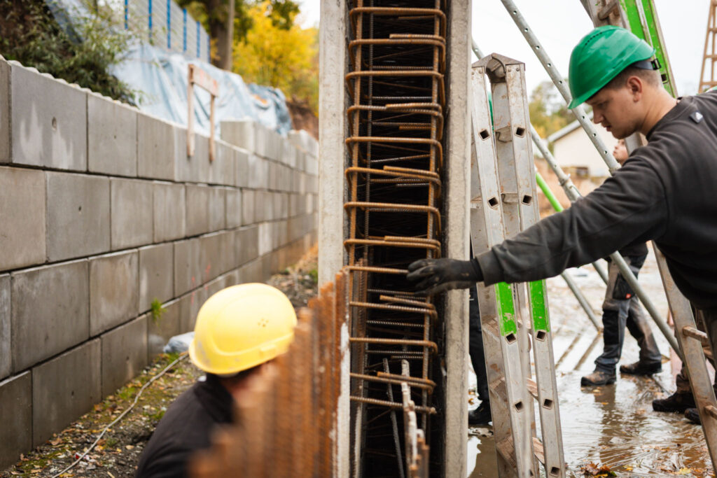 Bauarbeiter in Schutzkleidung inspiziert eine Bewehrung aus Stahlmatten in einer Säulenschalung auf einer Baustelle.