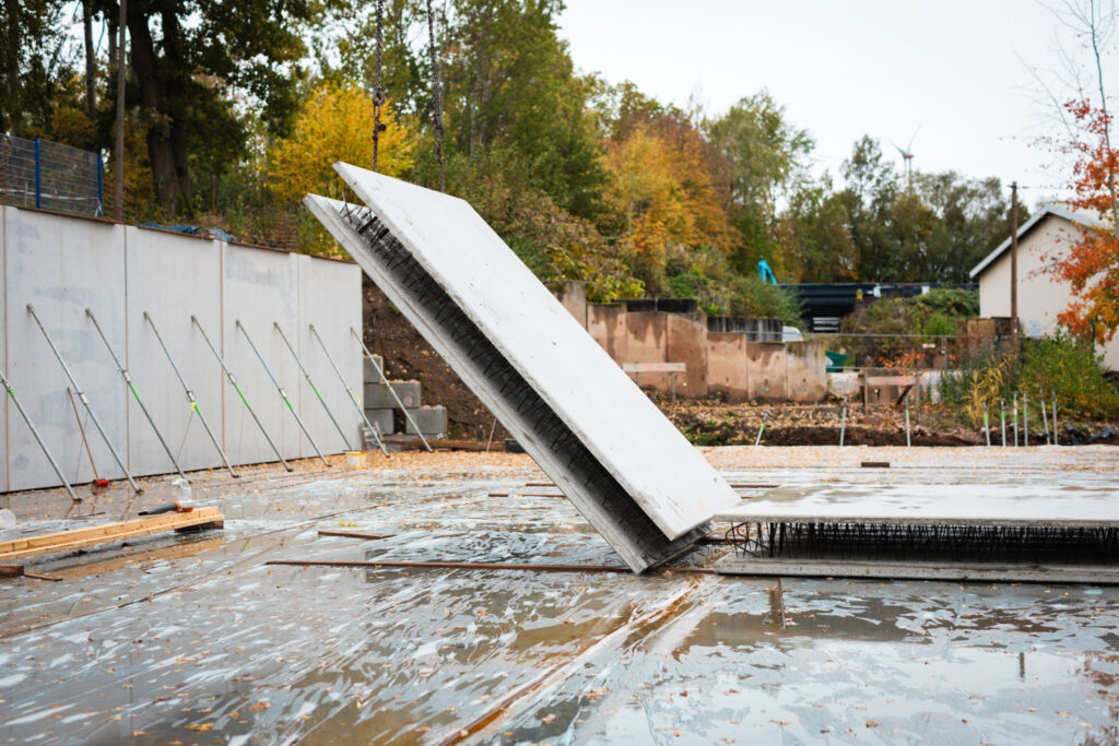 Zwei große, schräg stehende Betonfertigplatten mit Bewehrungsstäben auf einer Baustelle mit nassem Boden und Herbstbäumen im Hintergrund.