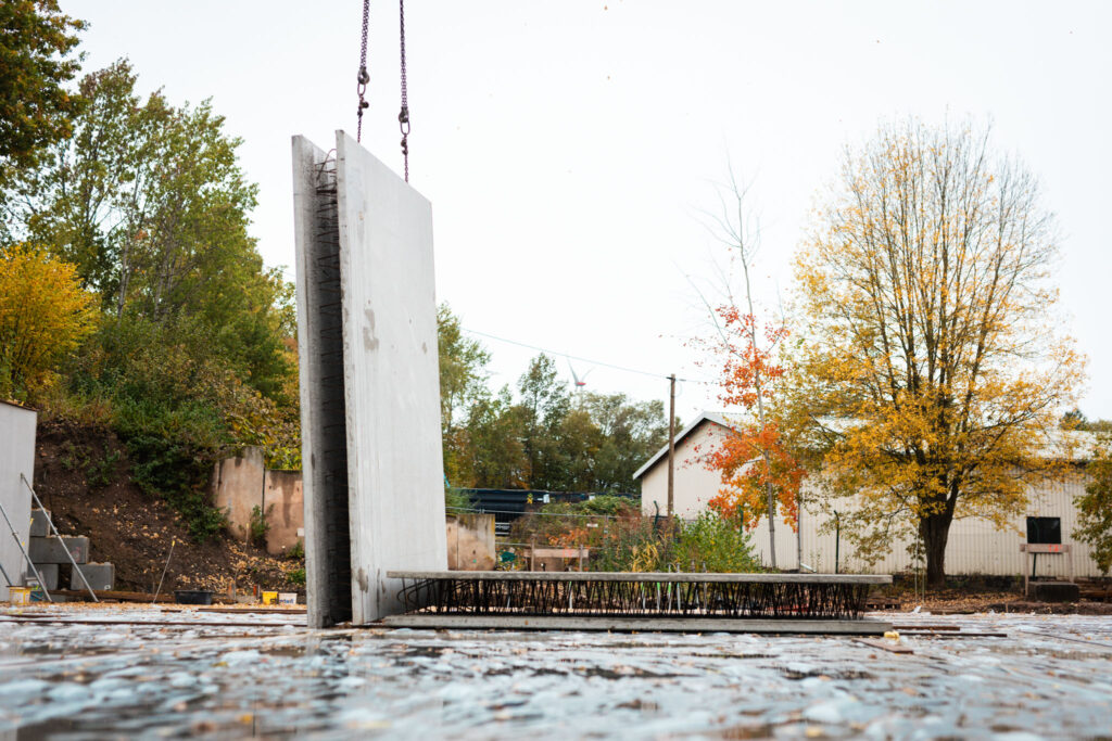 Zwei große Betonfertigteile stehen senkrecht und waagerecht auf einer nassen Baustellenfläche, umgeben von herbstlichen Bäumen und einem Gebäude.
