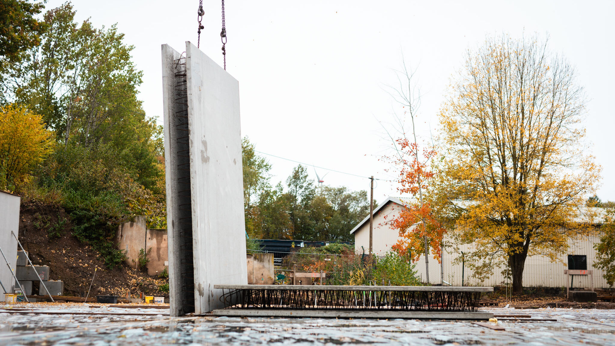 Zwei große Betonfertigteile stehen senkrecht und waagerecht auf einer nassen Baustellenfläche, umgeben von herbstlichen Bäumen und einem Gebäude.
