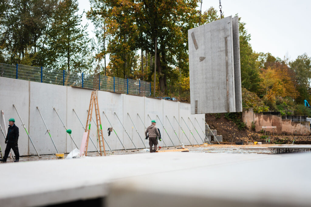 Ein Kran hebt eine große, rechteckige Betonwandplatte auf einer Baustelle, mit einem Bauarbeiter im Vordergrund und einer langen Stützmauer.