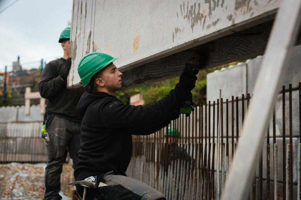 Bauarbeiter mit grünem Schutzhelm bearbeitet mit einem Winkelschleifer eine aufgehängte Betonfertigplatte auf einer Baustelle.