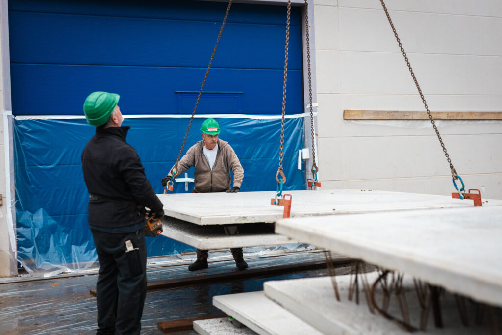 Zwei Bauarbeiter in Schutzhelmen heben mit einem Kran und Ketten eine Betonfertigplatte auf einer industriellen Baustelle ab.