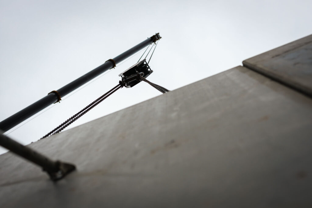 Untersicht einer Kranvorrichtung mit Kette, die eine Betonwand oder Fertigplatte auf einer Baustelle hochzieht.
