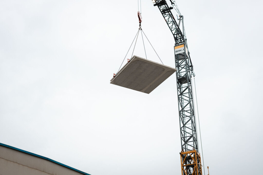 Ein hoher Bau- oder Turmkran hebt eine große rechteckige Betonfertigplatte gegen einen hellen, bewölkten Himmel.