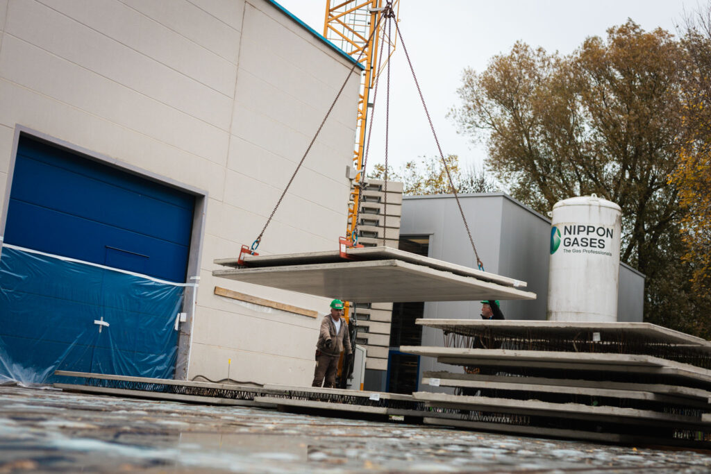 Zwei Bauarbeiter überwachen den Transport von gestapelten Betonfertigplatten, die von einem Kran vor einer Lagerhalle gehoben werden.