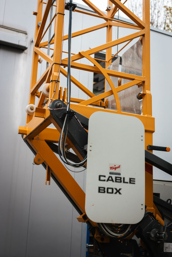 Detailaufnahme des gelben Fachwerks und einer weißen "Cable Box" Beschriftung am Fuß eines Krans auf einer Baustelle.