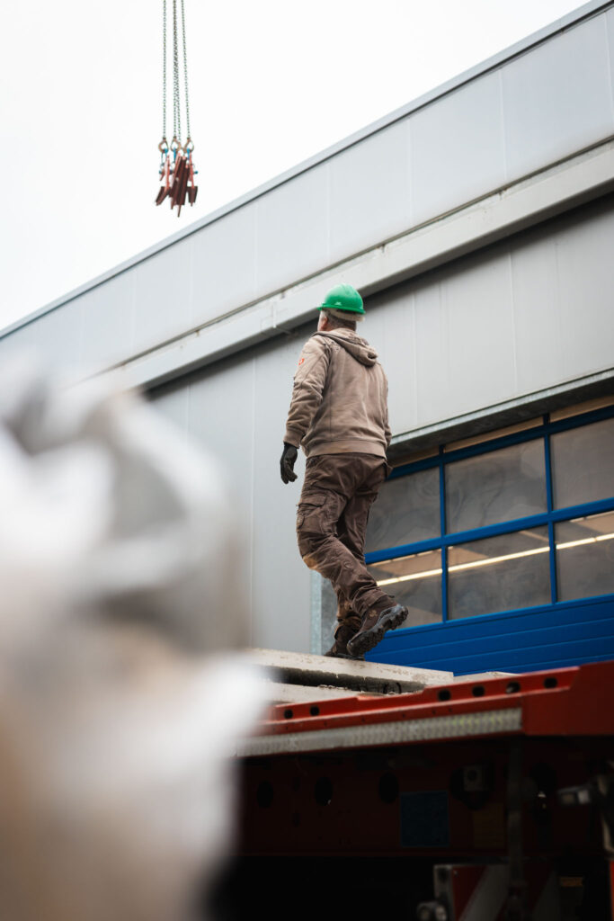 Bauarbeiter mit grünem Schutzhelm läuft über die Ladefläche eines Lastwagens vor einem Lagerhaus, während ein Kranhaken von oben herabhängt.