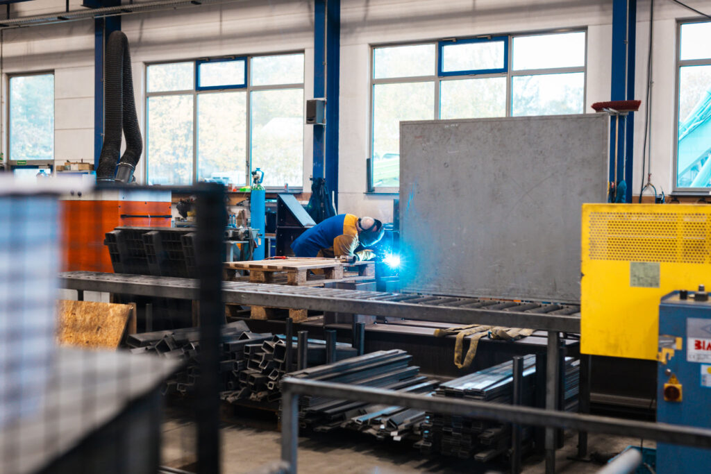 Gesamtansicht einer Werkstatthalle mit großen Fenstern. Ein Arbeiter in gelb-blauer Arbeitskleidung ist in der Mitte des Bildes bei Schweißarbeiten an einer erhöhten Metallstruktur zu sehen.
