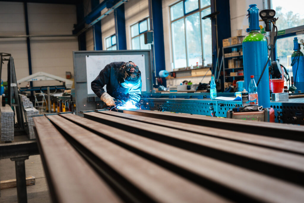 Ein Arbeiter in Schutzkleidung schweißt Metallträger in einer großen Lagerhalle oder Werkstatt. Die helle Flamme des Schweißbrenners und der Schutzhelm des Arbeiters sind deutlich sichtbar. Im Vordergrund liegen lange, dunkle Metallprofile.