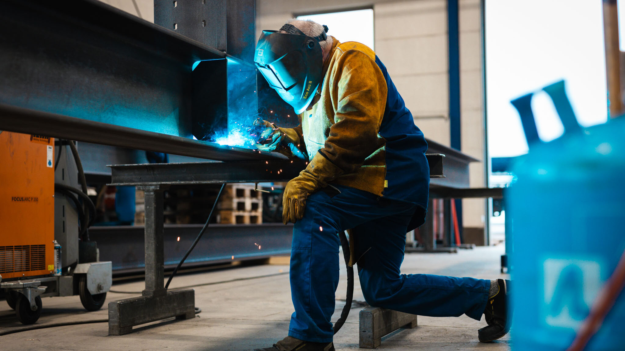 Ein Schweißer mit Schutzhelm kniet vor einem langen Stahlträger und schweißt die Nähte. Blaues Licht und Funken sprühen.
