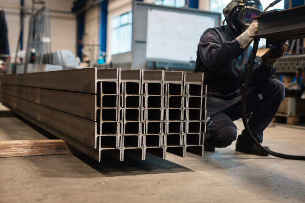 Nahaufnahme eines gestapelten Bündels von I- oder H-förmigen Stahlträgern. Im Hintergrund kniet ein Arbeiter in dunkler Kleidung und Schweißhelm, der einen Träger bearbeitet.