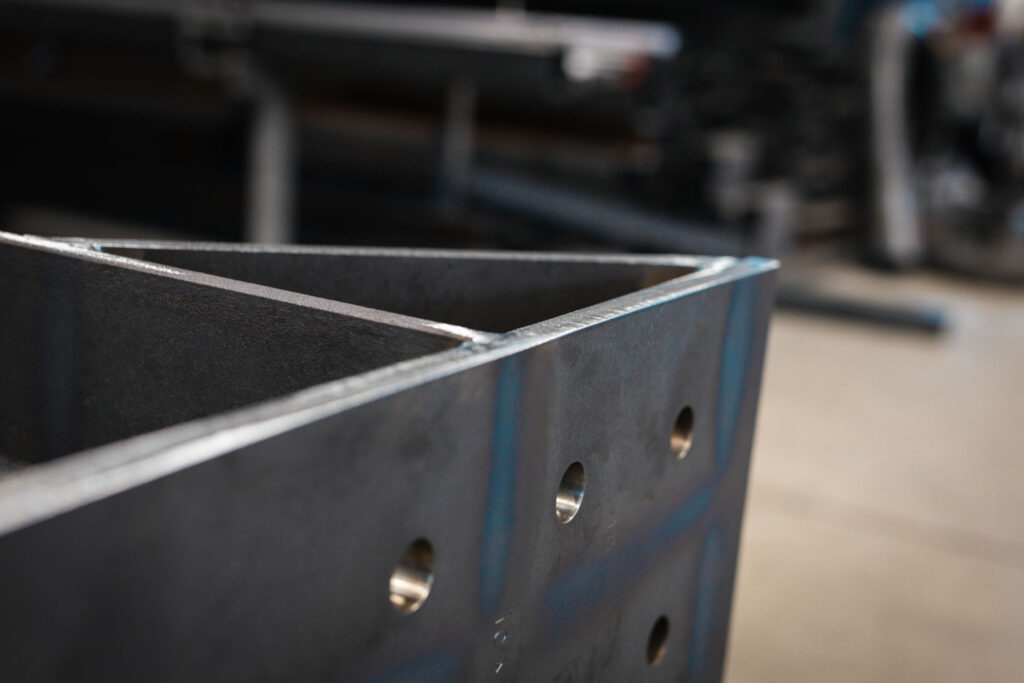 Detailaufnahme einer Ecke einer geschweißten Metallkonstruktion. Die Kanten sind sauber geschnitten und geschweißt. Man sieht mehrere runde Bohrlöcher in der Seite des Metalls.