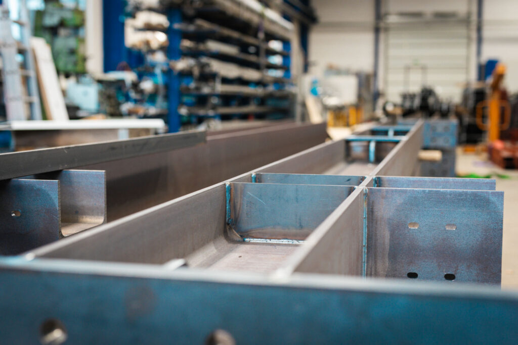 Nahaufnahme einer in der Entstehung begriffenen, geschweißten Metallstruktur, die aus langen Seitenwänden und rechtwinkligen Querstreben besteht. Die Oberfläche des Metalls ist roh und dunkel.