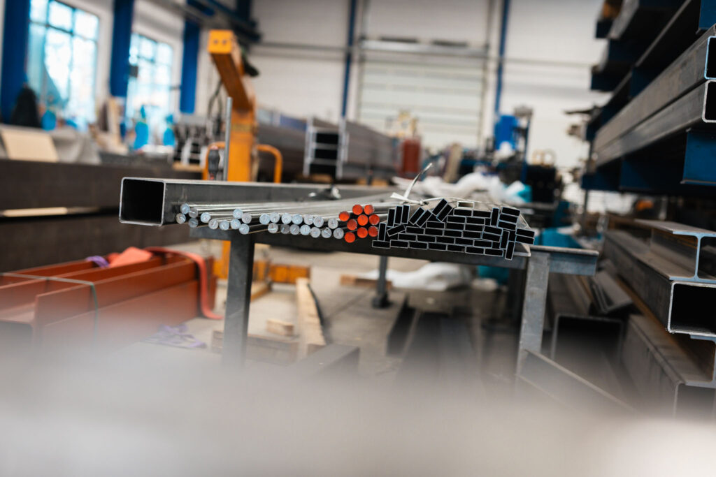Detailansicht eines Arbeitstisches in einer Werkstatt, auf dem ein Stapel quadratischer Hohlprofile und ein Bündel runder Metallstäbe liegt. Ein Ende der runden Stäbe ist mit orangefarbenem Kunststoff umwickelt.