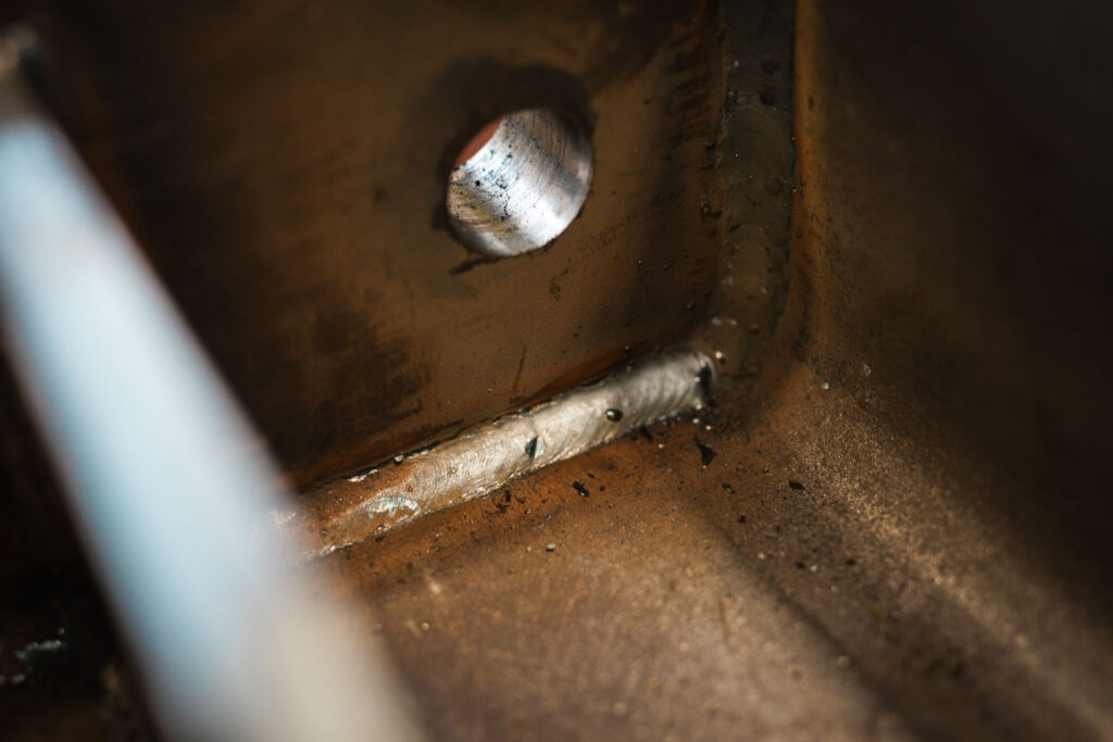 Extreme Nahaufnahme einer inneren Ecke eines Metallprofils, die eine deutliche, durchgehende Schweißnaht und ein großes, kreisrundes Bohrloch zeigt. Die Oberfläche ist braun-rostig und rau.