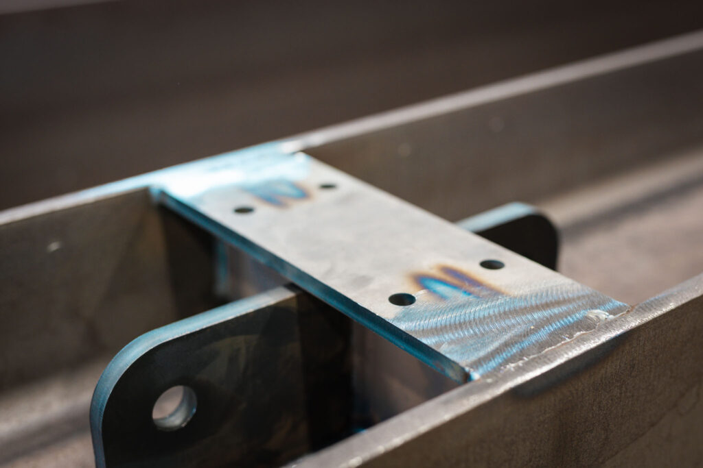 Nahaufnahme einer geschweißten Verbindung, bei der eine flache Metallplatte mit Bohrlöchern auf einen größeren Metallträger montiert ist. Die Schweißnähte zeigen die typischen blauen und braunen Spiegelungen.