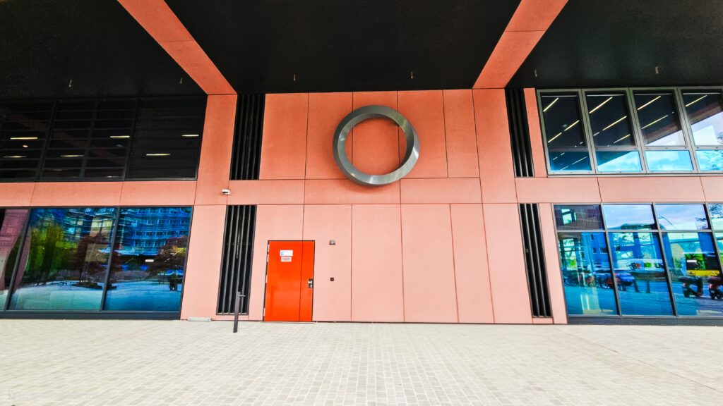 Frontale Ansicht des Eingangs eines Geschäftsgebäudes. Die Fassade ist in sanftem Korallenrot gehalten, flankiert von großen, reflektierenden Fensterflächen. In der Mitte ist ein großes, stahlfarbenes, in sich verdrehtes Ring-Schild oder Logo befestigt, darunter eine leuchtend rote Tür.