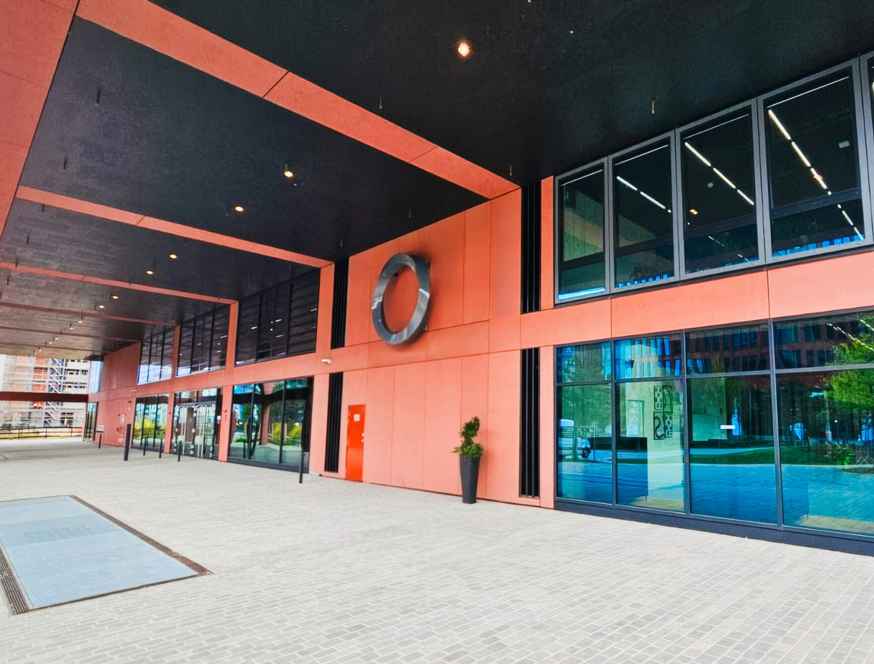 Eingangsbereich eines modernen Bürogebäudes in Hamburg mit schwarzen Decken und korallenroten Fassadenplatten. Ein großes, silbernes, verdrehtes ringförmiges Logo (Edge Logo) ist an der Wand montiert. Unten links steht "IDEEN AUS METALL EDGE LOGO // HAMBURG".