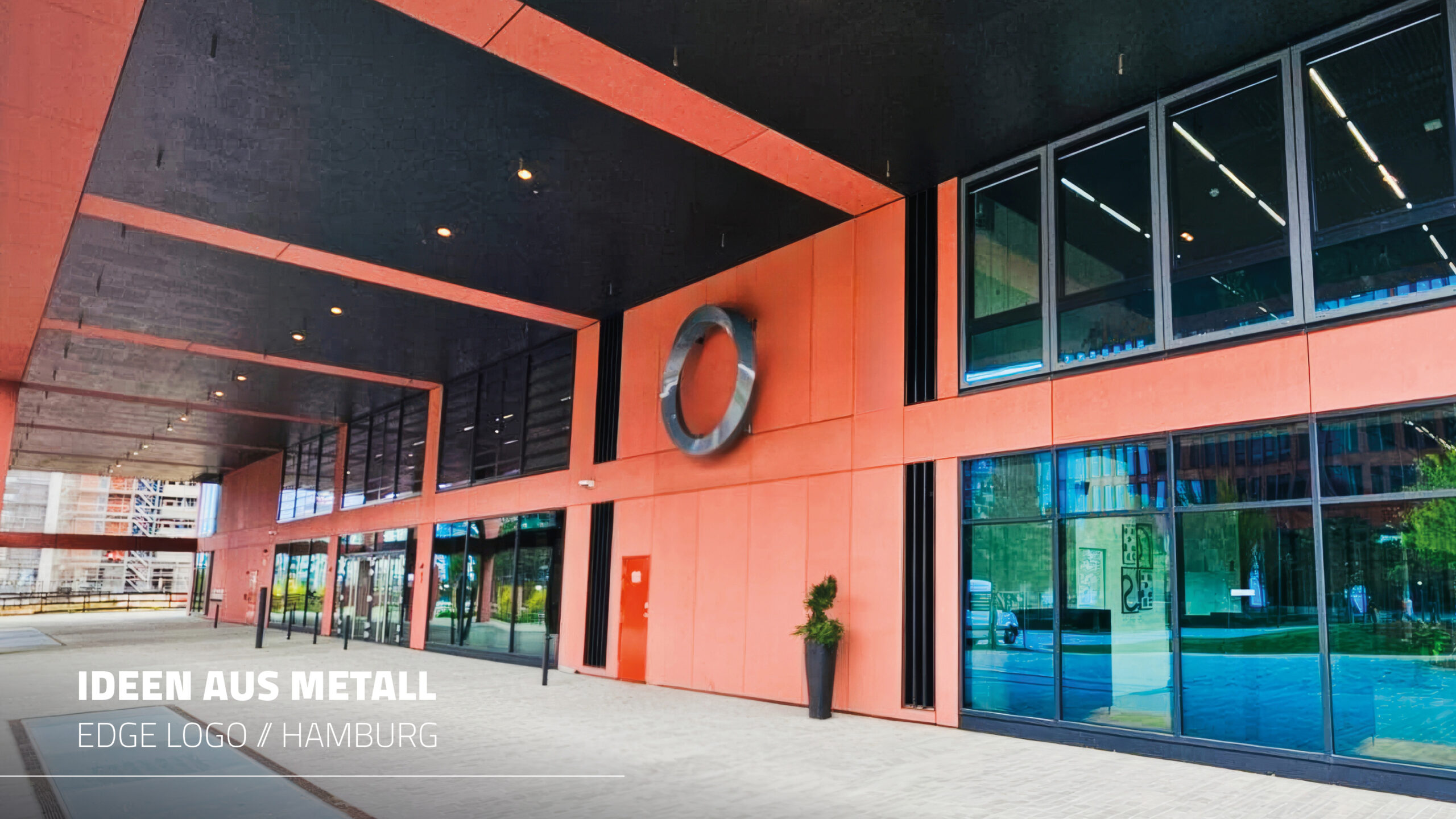 Eingangsbereich eines modernen Bürogebäudes in Hamburg mit schwarzen Decken und korallenroten Fassadenplatten. Ein großes, silbernes, verdrehtes ringförmiges Logo (Edge Logo) ist an der Wand montiert. Unten links steht "IDEEN AUS METALL EDGE LOGO // HAMBURG".