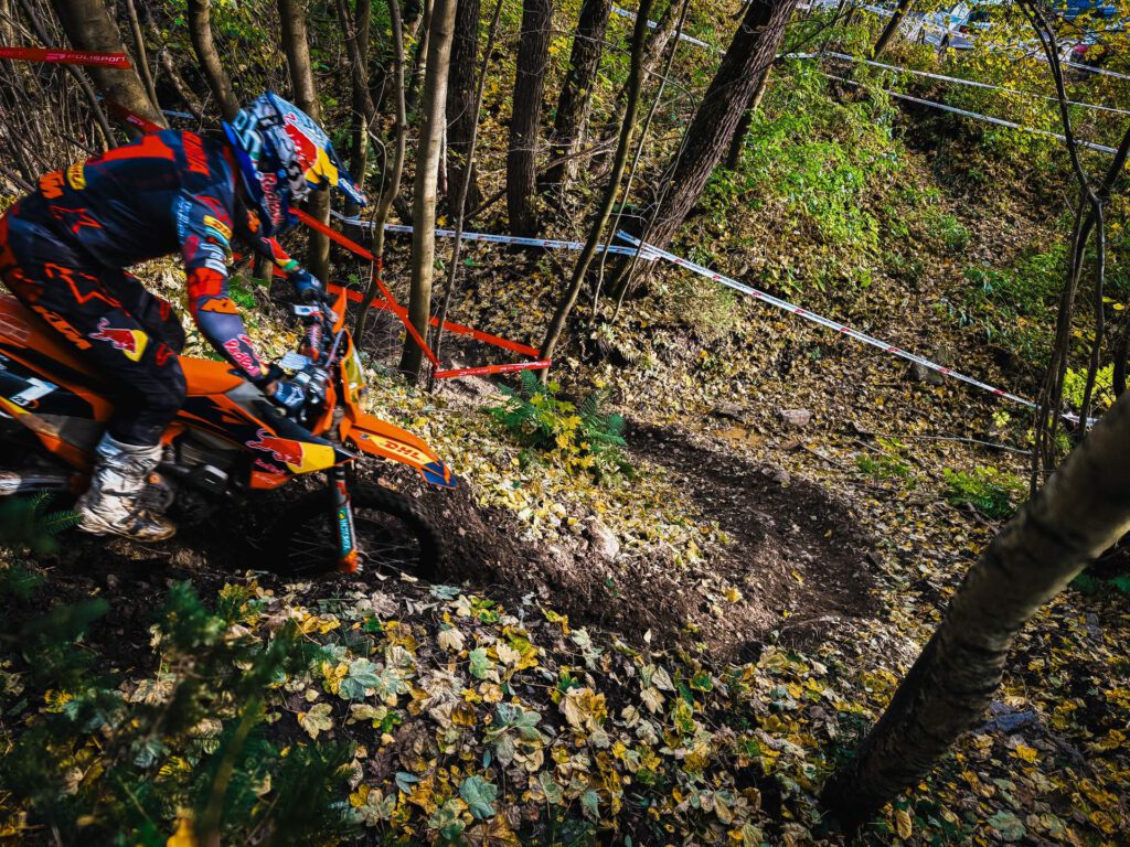 Dynamische Aufnahme eines Enduro-Rennfahrers in orange-schwarzer Red Bull-Ausrüstung, der mit seiner KTM durch eine schlammige, von Bäumen gesäumte Waldpassage fährt. Der Boden ist bedeckt mit Herbstlaub und matschigen Spuren.