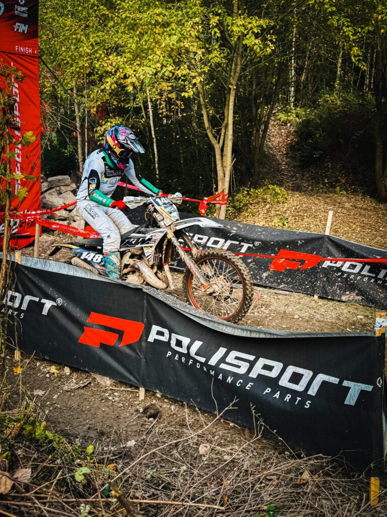 Enduro-Fahrerin mit der Startnummer 148 in weißer Kleidung fährt mit ihrem Motorrad über eine Sektion mit großen, schwarzen POLISPORT-Bannern. Die Strecke ist schlammig und befindet sich in einem Waldstück, nahe einem Finish-Tor. (
