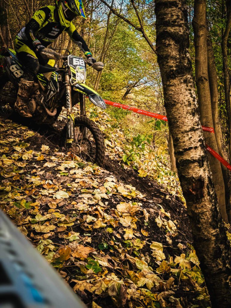 Motocross-Fahrer mit der Nummer 68 auf einem gelb-schwarzen Motorrad fährt einen rutschigen, mit Herbstlaub bedeckten Waldhang hinauf. Die Fahrbahn ist matschig und von Bäumen gesäumt.