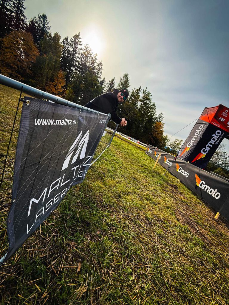Zuschauer in schwarzer Kleidung lehnt an einem Absperrband am Zielbereich einer Offroad-Veranstaltung. Im Vordergrund ein Banner des Sponsors MALTITZ mit der URL www.maltitz.de. Der Hintergrund zeigt Wald und Sonnenschein.