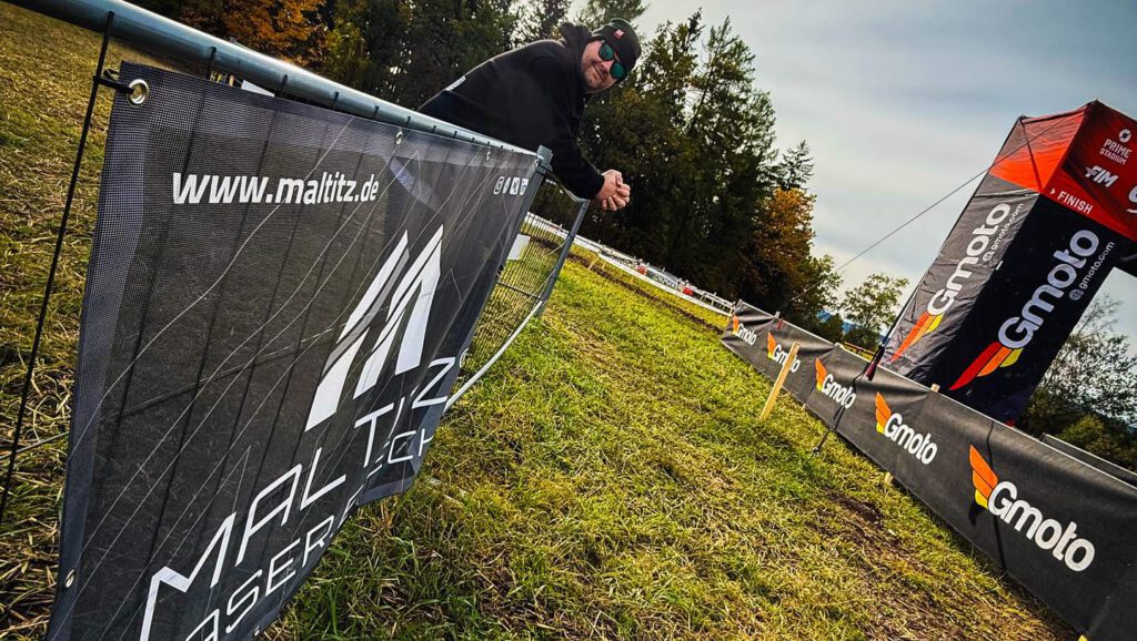 Zuschauer in schwarzer Kleidung lehnt an einem Absperrband am Zielbereich einer Offroad-Veranstaltung. Im Vordergrund ein Banner des Sponsors MALTITZ mit der URL www.maltitz.de. Der Hintergrund zeigt Wald und Sonnenschein.