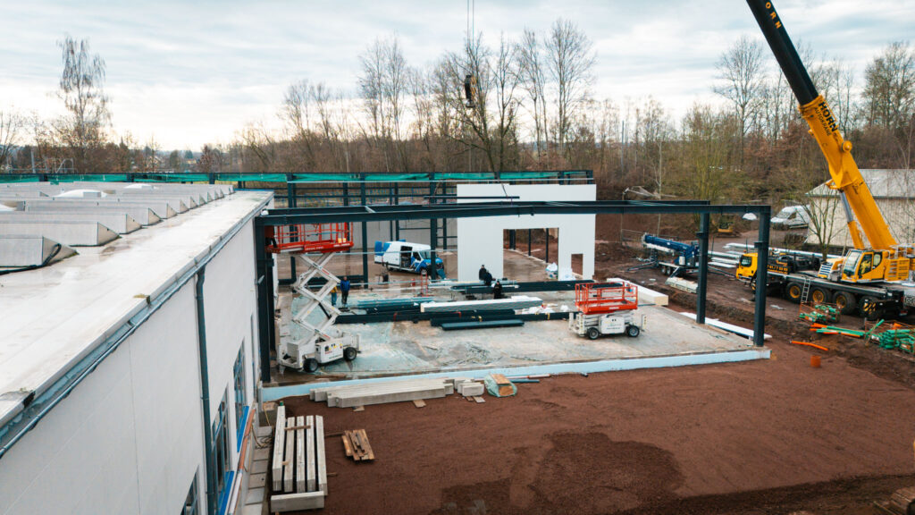Eine Gesamtansicht der Baustelle mit dem Stahlgerüst und den weißen Wandelementen. Zwei Arbeiter sind auf dem Fundament im Zentrum der Konstruktion zu sehen, umgeben von Baumaschinen wie Hubarbeitsbühnen und Material. Der Boden ist eine Mischung aus Erde und Beton.