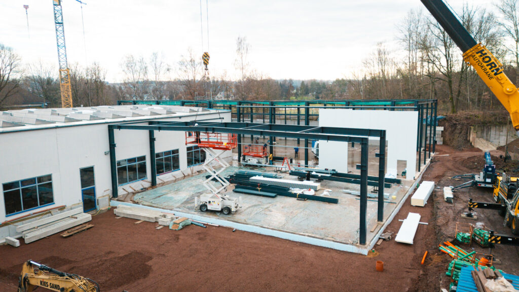 Eine Panoramaaufnahme der Baustelle von oben oder aus der Ferne. Die Stahlrahmenkonstruktion ist teilweise mit weißen Fassadenelementen verkleidet. Die Betonbodenplatte ist erkennbar, und ein Kran ist im Hintergrund sichtbar.