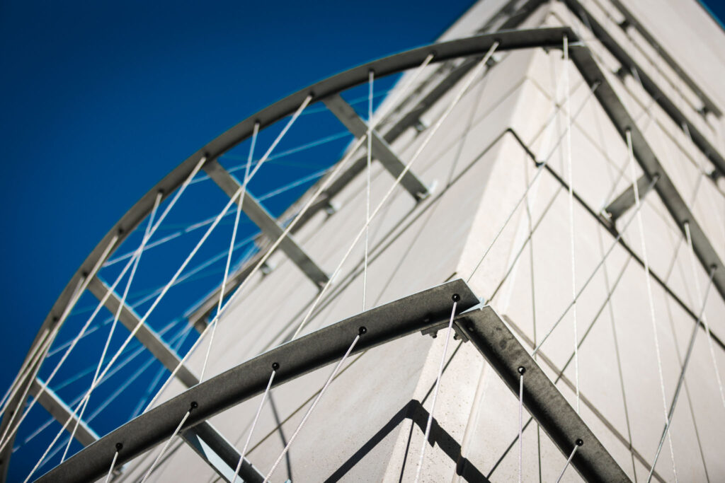 Nahaufnahme einer Eckkonstruktion mit einem runden Metallgeländer, das wahrscheinlich zu einem der Balkone der hellen Fassade gehört.