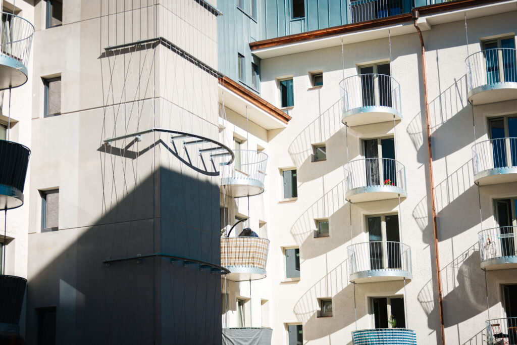 Reihe von kleinen, runden Balkonen mit schlichten Metallgeländern an einer hellen Wohnhausfassade, die lange Schatten werfen.