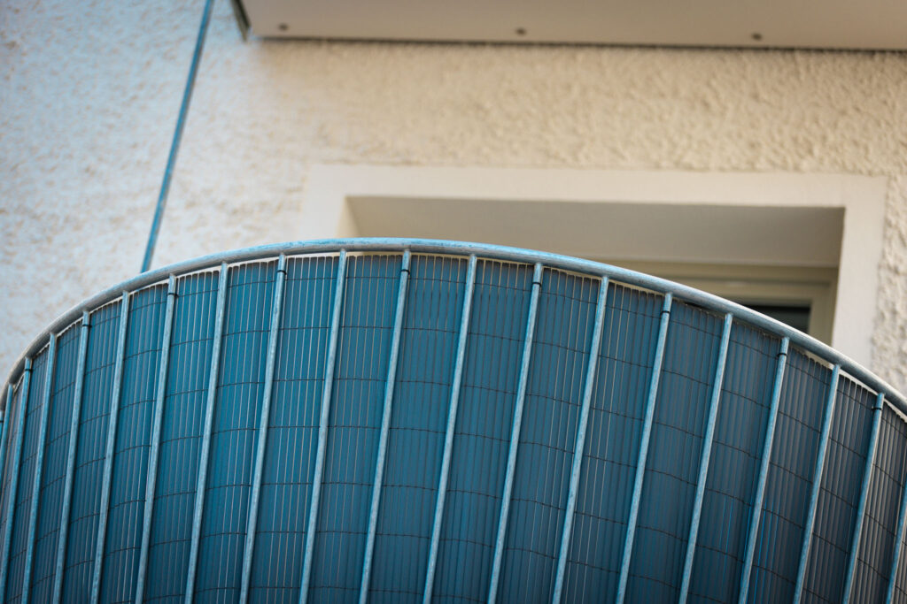 Nahaufnahme des runden Balkongeländers, das aus vertikalen Metallstäben und einem blickdichten Sichtschutz an der Innenseite besteht.